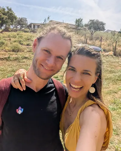 Lexmy and Simon, a selfie in a sunny field, with trees and a house on the slope behind them.